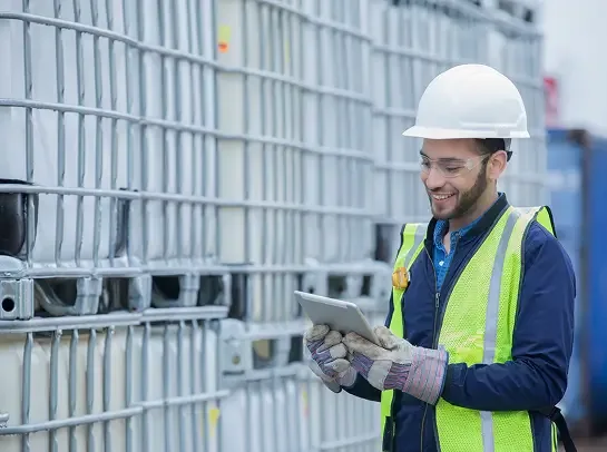 A smiling person wearing a hard hat, safety glasses, and a reflective vest looks at a tablet in front of stacked industrial storage tanks.