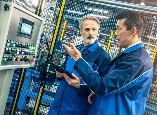 Two workers in blue uniforms discuss a machinery control panel inside a factory; one holds a tablet and gestures toward the illuminated buttons.