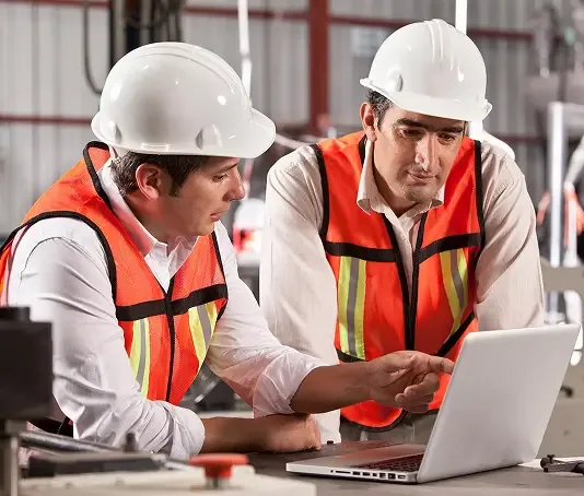 Two individuals wearing hard hats and safety vests review information on a laptop computer in an industrial setting.