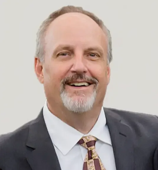 A smiling man with a graying goatee, wearing a dark suit jacket, white shirt, and a patterned tie.