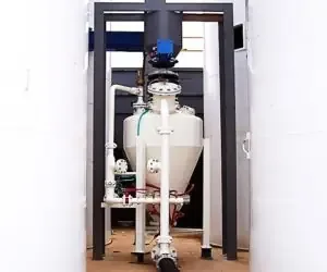 White pneumatic blow tank on a black steel frame between silos, with flanged piping and valves.