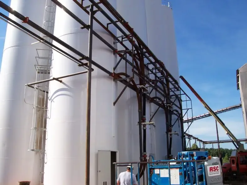 Tall white outdoor silos with piping and ladder; worker and blue aerial lift in the foreground.