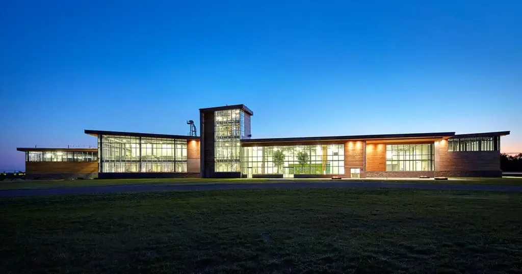 Modern glass-and-wood distillery building glowing at dusk across a green lawn.