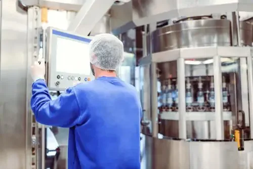 Worker in a blue shirt and hairnet monitors a stainless steel control panel beside a rotary bottling machine with clear bottles.