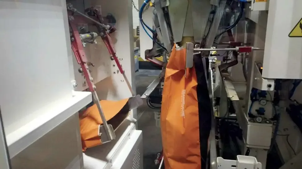 Orange feed bags held in metal grippers inside an automated bagging machine.