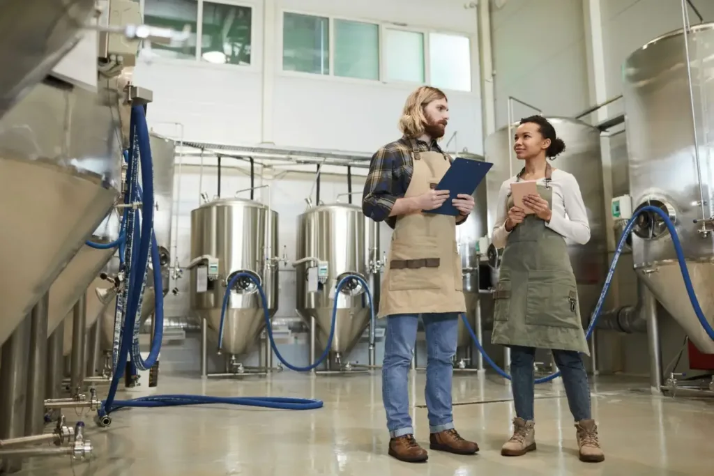 Two workers in aprons discuss production beside conical stainless steel tanks with blue hoses on the brewery floor.
