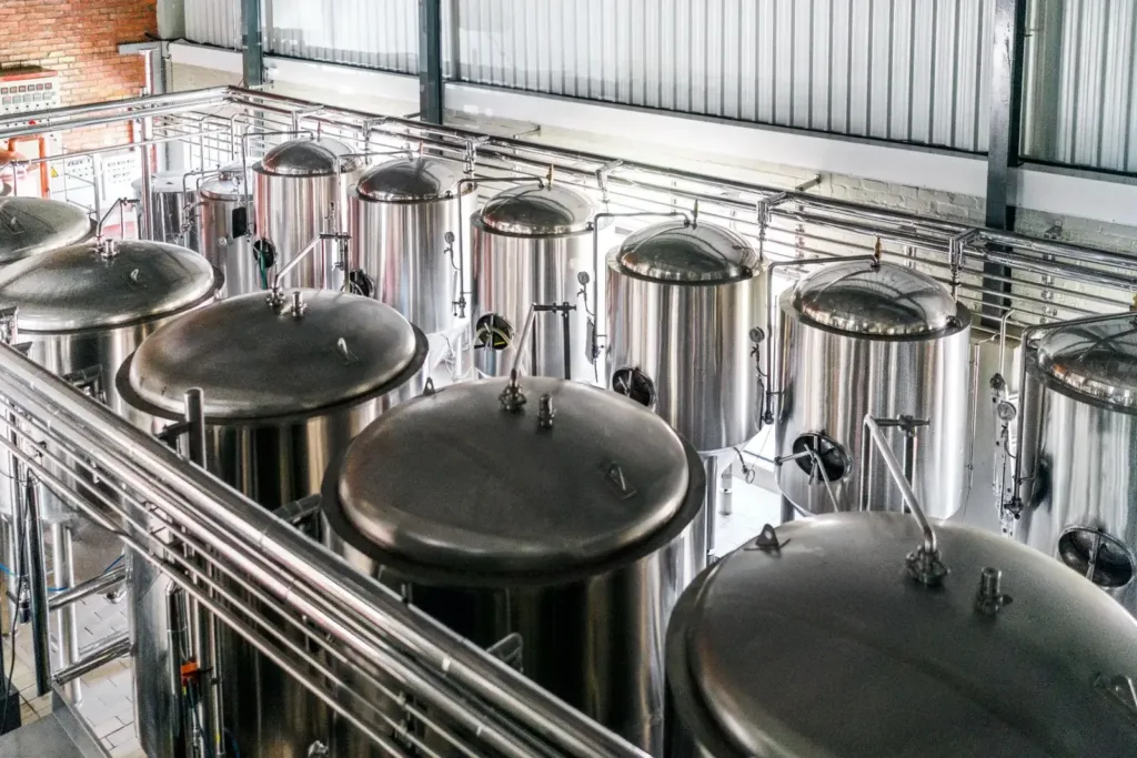 Rows of polished stainless steel fermentation tanks fill a bright brewery floor beneath overhead piping and walkways.