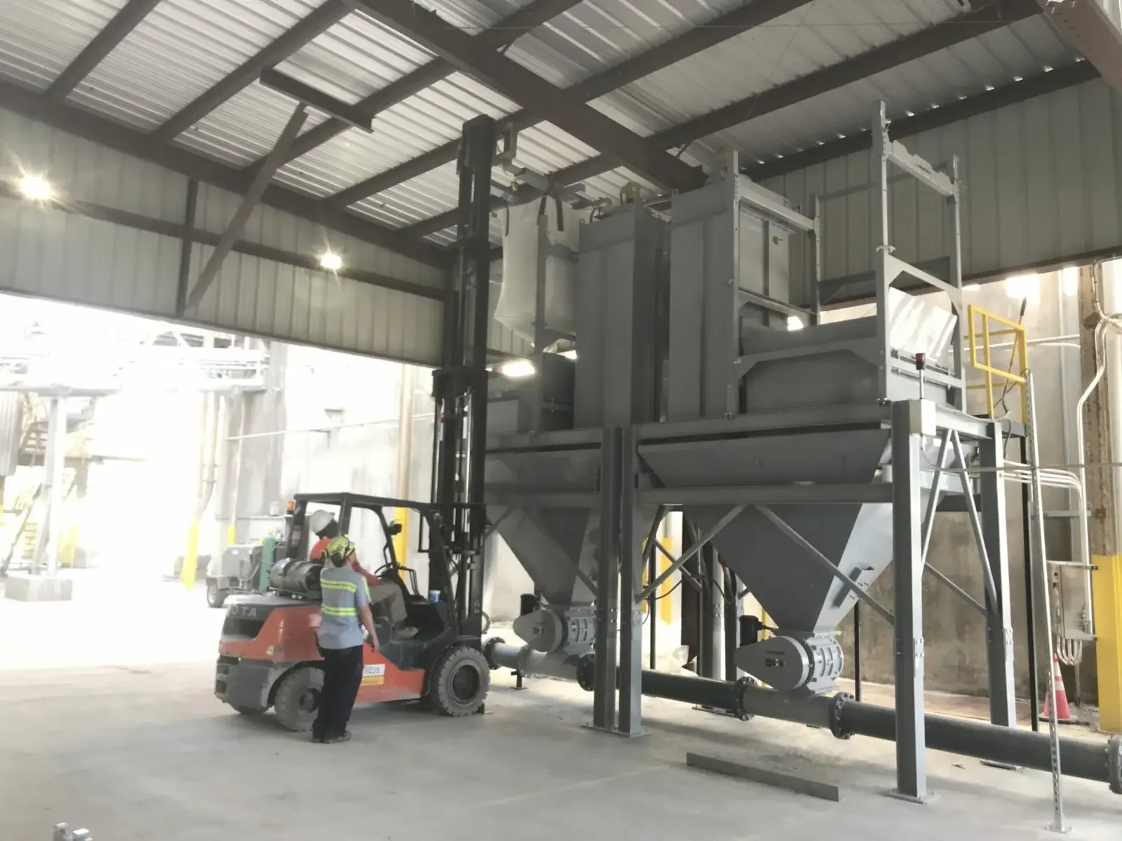 Forklift lifts a bulk bag into a gray industrial hopper system inside a warehouse.