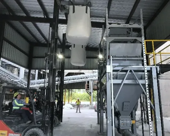 Forklift operator positions white bulk bags for unloading into a gray industrial hopper.