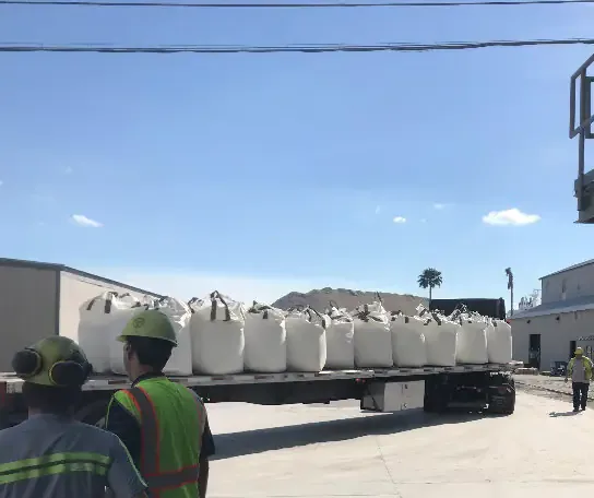 Flatbed truck loaded with white bulk bags at a facility, workers in safety gear nearby.