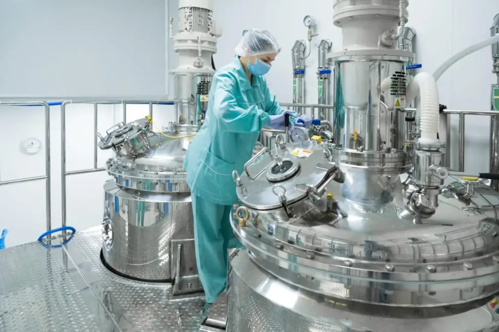 Cleanroom worker in a blue suit, mask, and gloves operates polished stainless steel reactors on a diamond-plate floor.