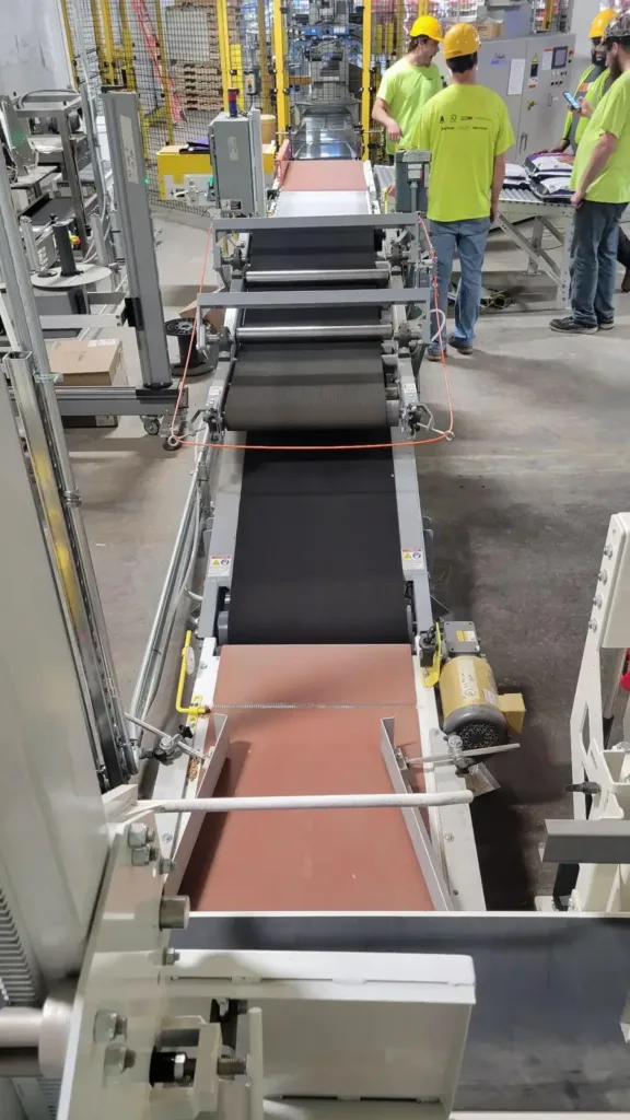 Workers in yellow shirts and hard hats stand along a newly installed multi-section conveyor line.