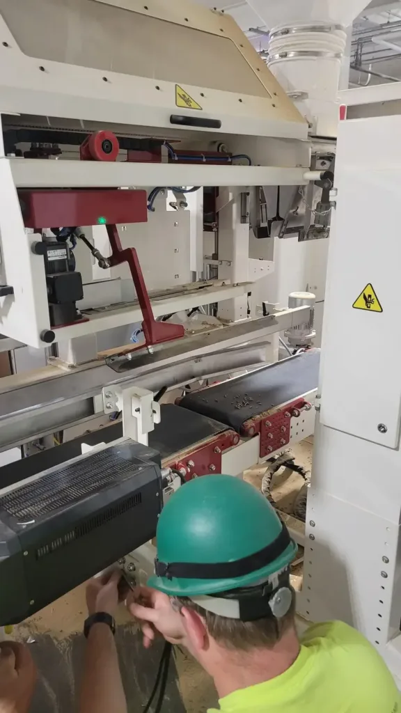 Technician in a green hard hat adjusts a component beneath a white bagging machine's conveyor decks.
