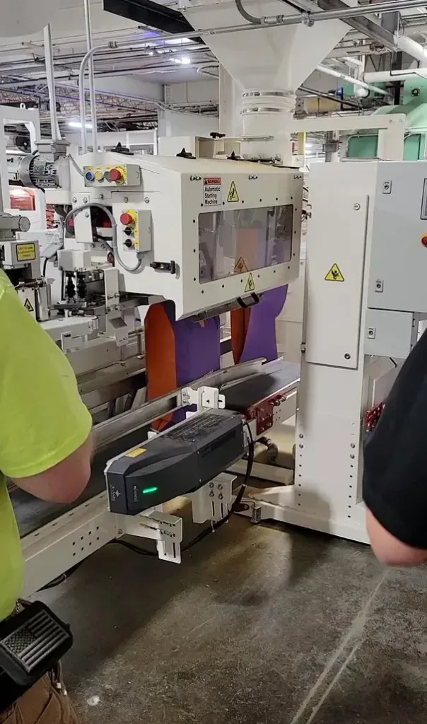 White automated bagger fills purple and orange bags on a black conveyor with control buttons and a hopper above.