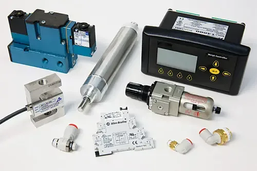 Assorted automation parts on a white surface: pneumatic valve, cylinder, weigh controller, load cell, filter regulator, and fittings.