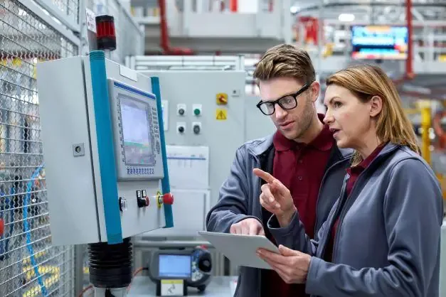 Two colleagues review a tablet beside an HMI control panel on a caged machine in a modern plant.