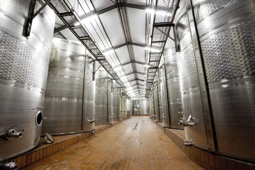 Long aisle between rows of tall stainless steel fermentation tanks with dimpled jackets under a high industrial ceiling.