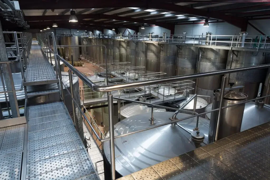 Elevated metal catwalk overlooks dozens of tall stainless steel storage tanks in a large beverage processing warehouse.