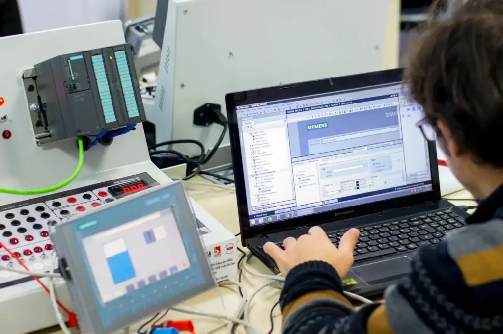Engineer programs Siemens TIA Portal on a laptop beside a PLC training panel and SIMATIC HMI touchscreen.