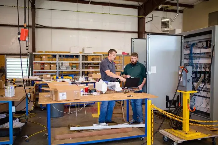Two technicians review a laptop at a workbench beside an open electrical control cabinet in a fab shop.