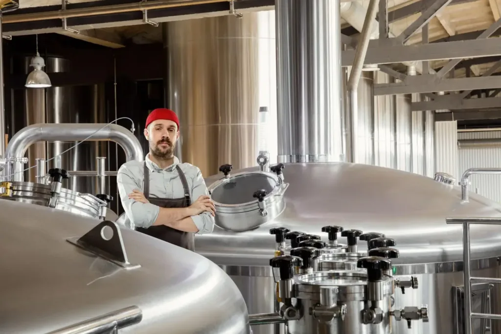 Bearded worker in a red beanie and apron stands arms crossed among large stainless steel tanks in a production plant.