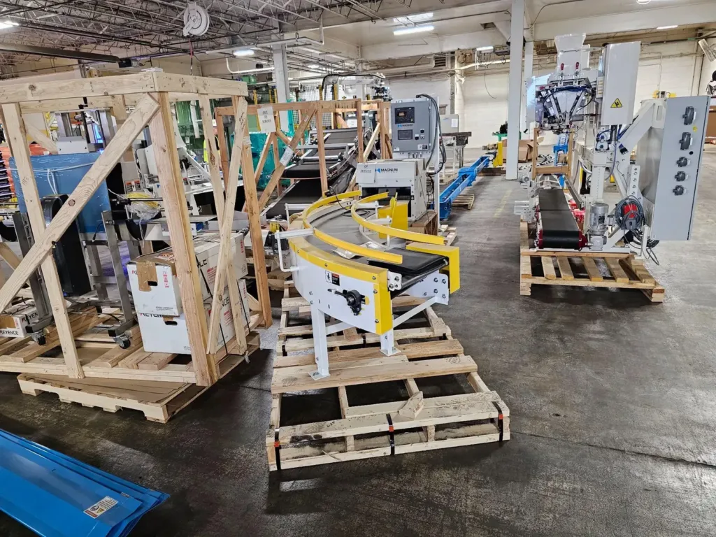 Curved yellow-guarded conveyor and crated equipment staged on wooden pallets in a warehouse.