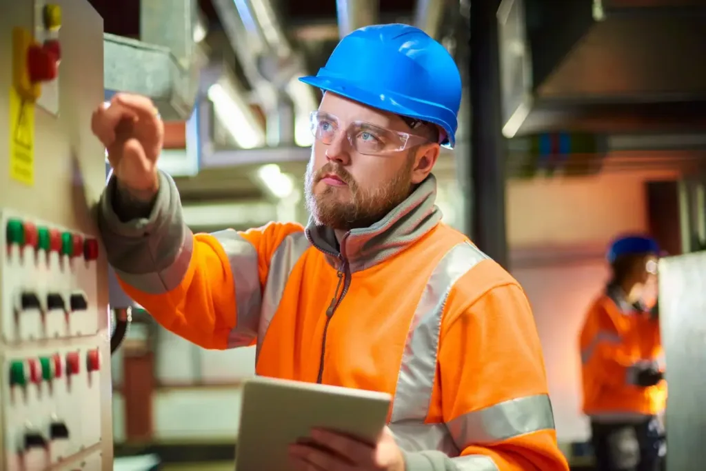 Technician in a blue hard hat and orange hi-vis jacket holds a tablet while adjusting a control panel.