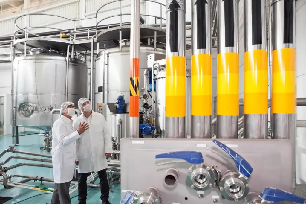 Two technicians in lab coats, hairnets, and masks discuss stainless steel tanks and color-banded process piping.