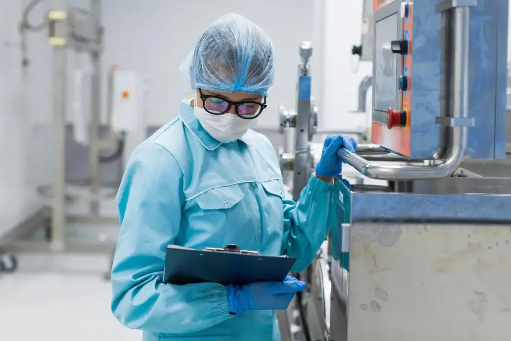 Gloved technician in a blue coat, hairnet, and mask reviews a clipboard beside a stainless steel machine control panel.