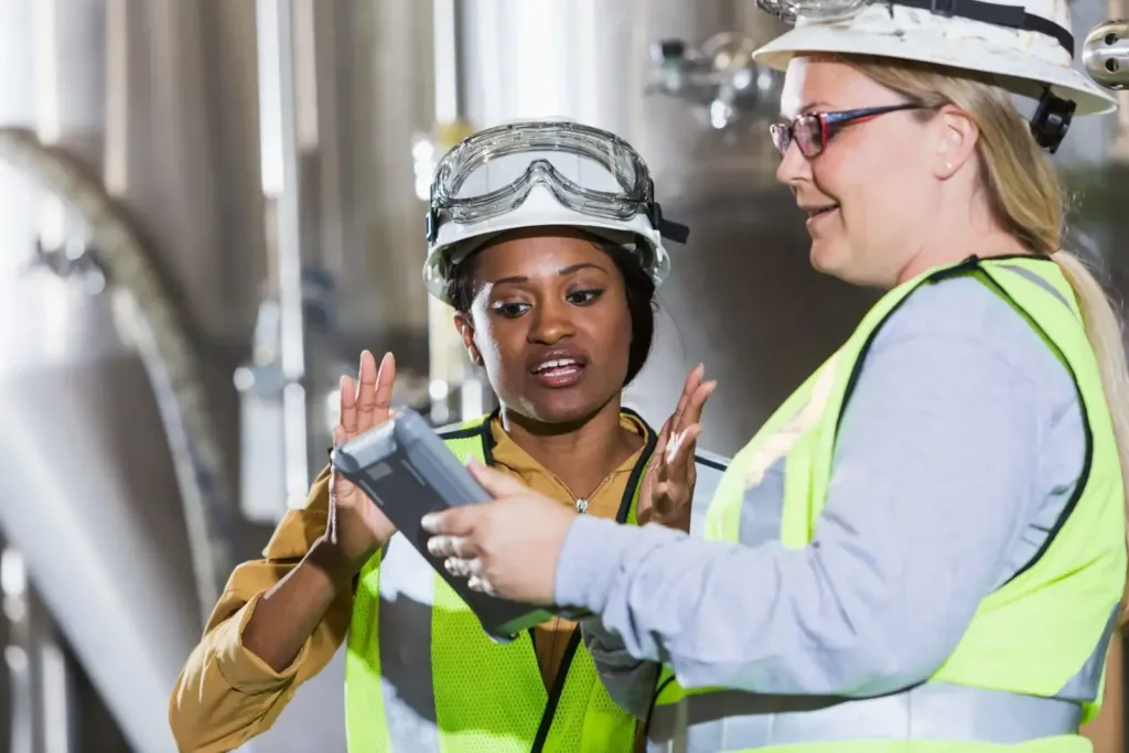 Two women in hard hats and high-vis vests review a rugged tablet beside stainless steel process tanks.