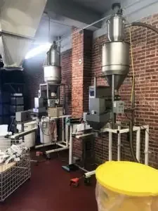 Two stainless steel coffee bean loaders on white frames against an exposed red brick wall.