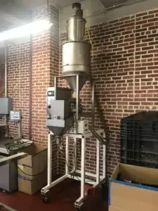 Vertical stainless steel hopper and dispensing system on a white mobile frame against a brick wall.
