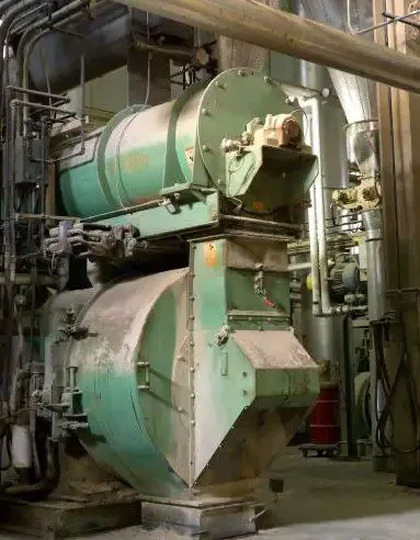 Dust-covered green industrial mill with a cylindrical feeder and hopper sits among plant piping and conduits.