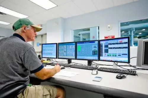 Operator in a green cap takes notes at a control-room desk with four monitors of process data.