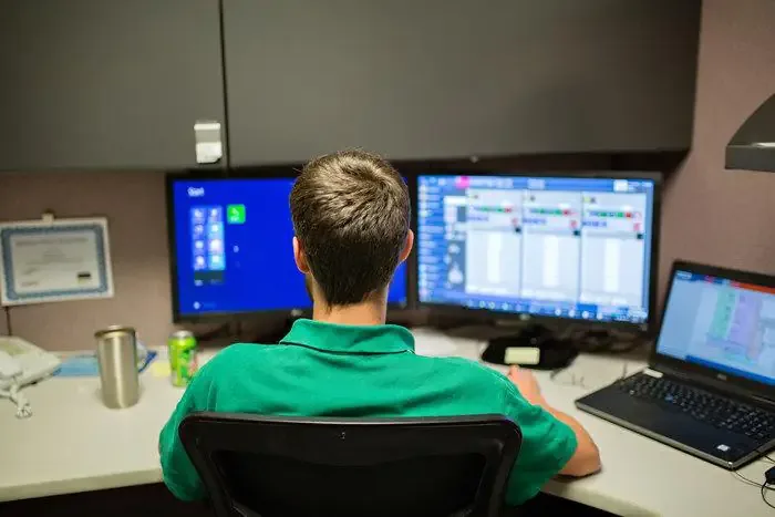 Engineer in a green polo works at a cubicle with dual monitors and a laptop showing production schedules.