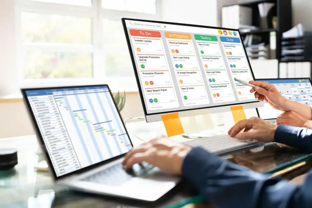 Hands type on a laptop Gantt chart while a monitor shows a color-coded Kanban board of project tasks.