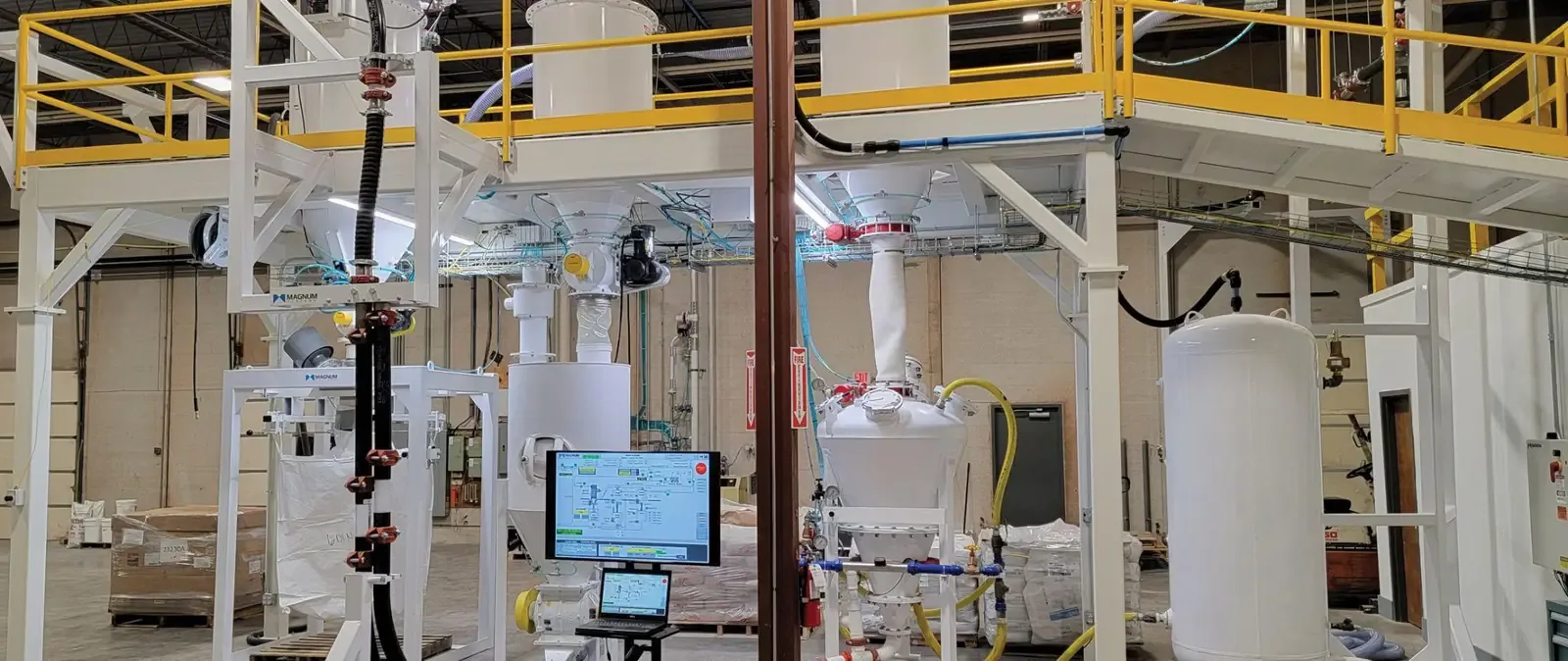 Magnum test lab with a bulk bag unloader, receivers, and pressure vessel beneath a yellow-railed mezzanine, HMI monitor in front.