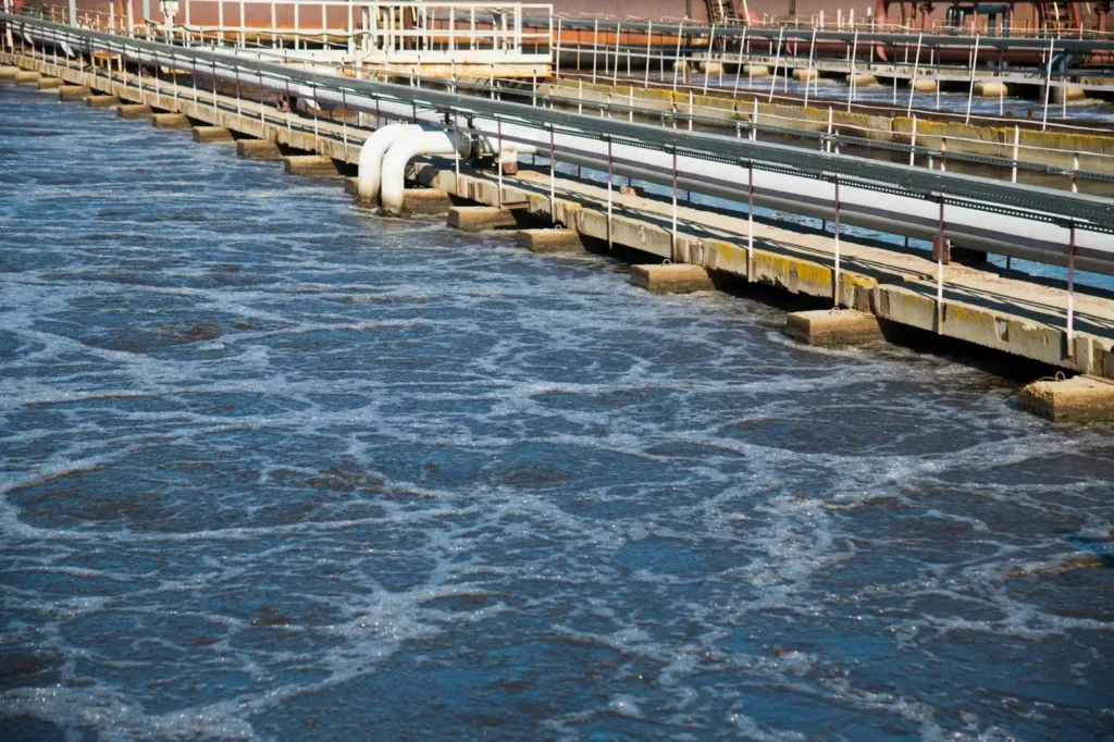 Rectangular aeration basin covered in white foam with a concrete walkway, railings, and white pipes overhead.