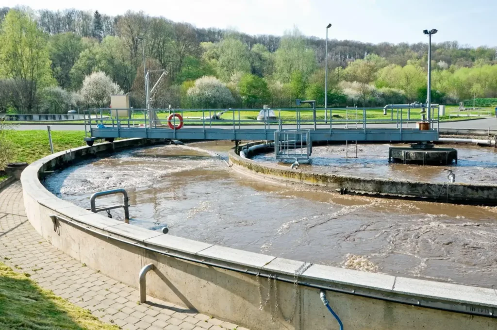 Circular outdoor aeration tank with churning brown water and a metal bridge walkway against a wooded hillside.