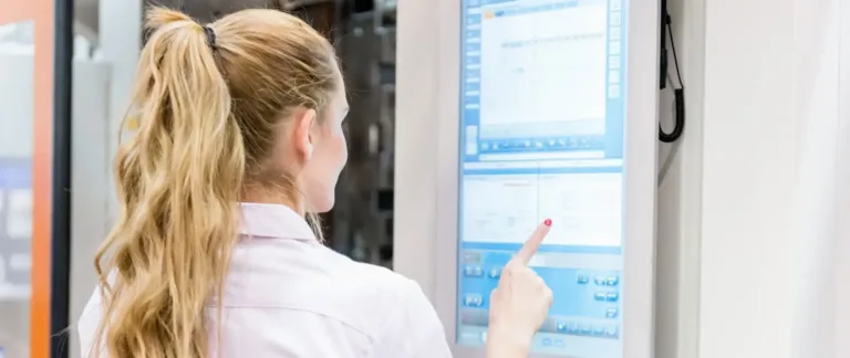 An operator works at a workstation with four computer monitors displaying control panel interfaces and data dashboards in a monitoring room.